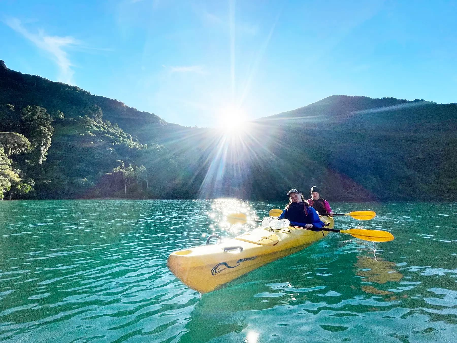 Two people paddling a tandem sea kayak on calm turquoise water as the sun rises over bush-clad hills