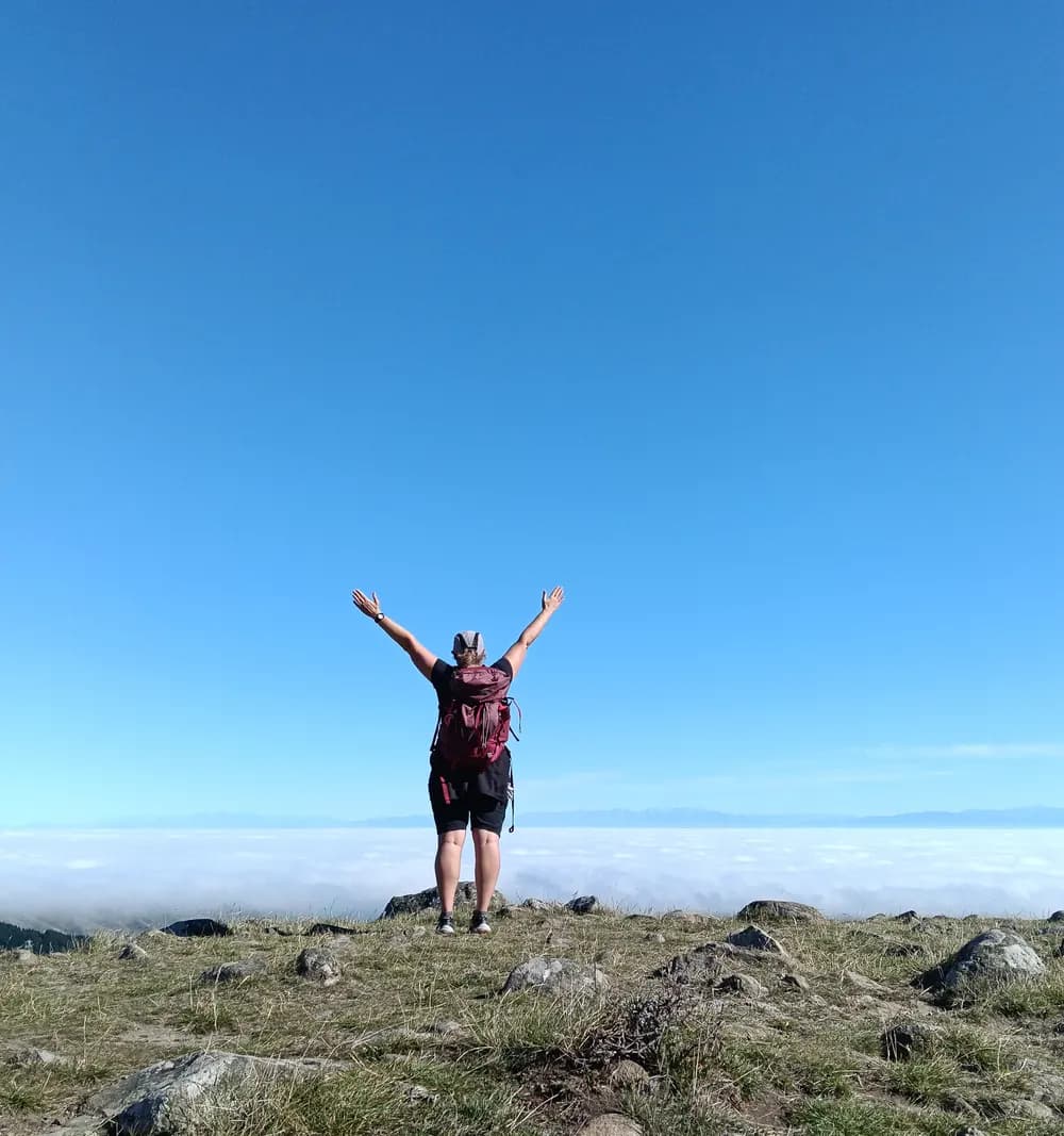 A person standing on a tussock ridge with their arms raised, looking out over a sea of cloud